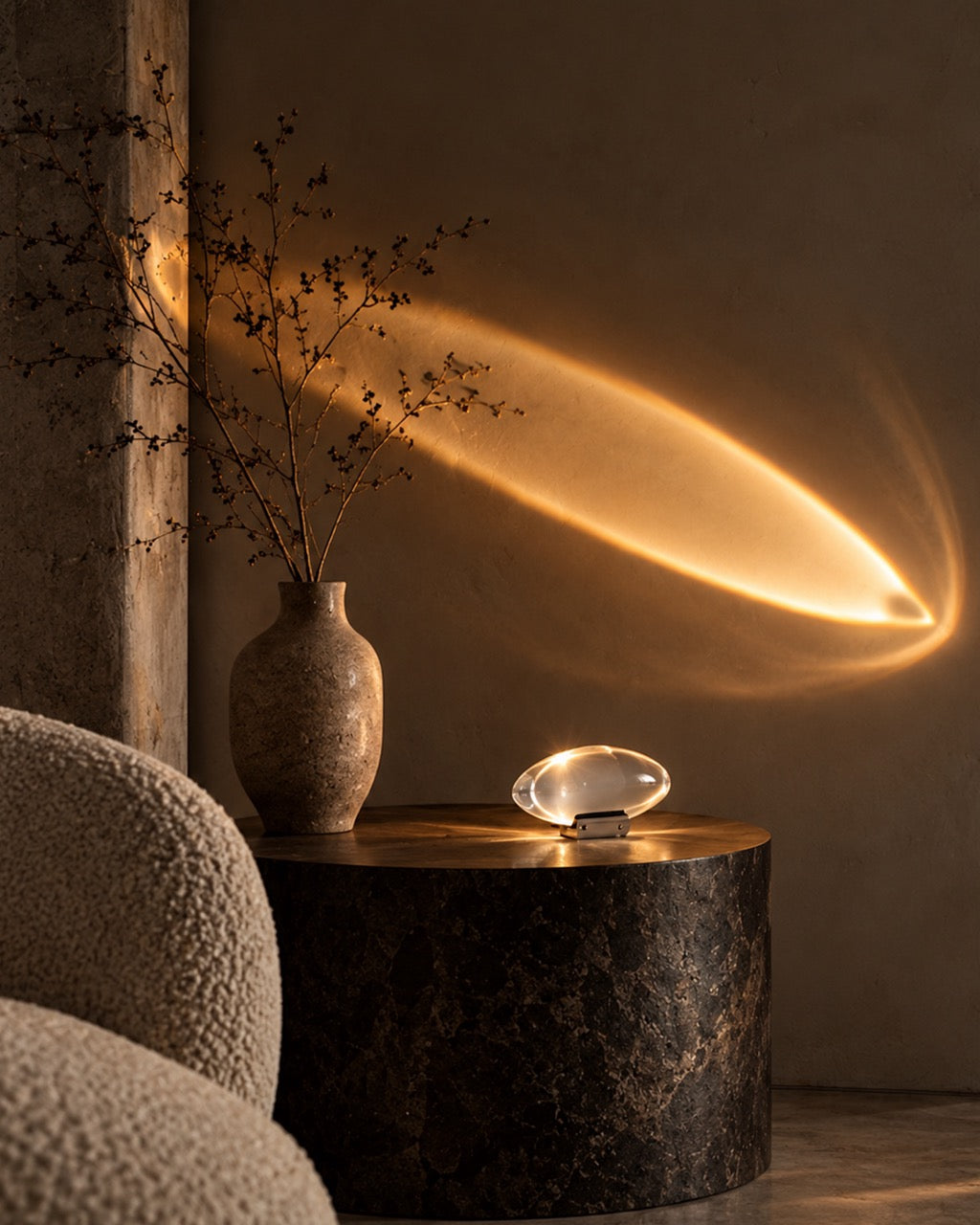 A modern oval table lamp with a clear glass shade and metallic base sits on a dark wood side table in a warm, moody room. The lamp casts a dramatic golden light onto the textured beige wall, creating a glowing streak.
