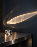 A clear glass or acrylic decorative object sits on a dark metallic tabletop, casting a warm glowing beam across a dark wall. The scene is photographed in a moody interior with strong reflections and no people or animals present.