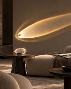 A modern living room scene with a warm glowing oval light fixture mounted on a beige wall, casting a soft golden light across the space. A textured beige pouf, dark wood side tables, and a glass orb lamp are visible in the room.