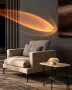 A modern beige upholstered armchair with matching cushions in a styled interior, next to a small round side table with a glowing sculptural lamp. The background features a dark gray wall with a large abstract light projection and a dark framed artwork.