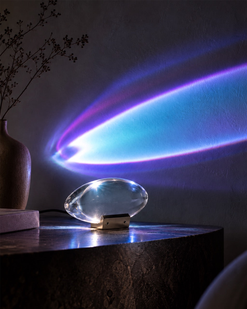 A clear oval-shaped LED light projector lamp sitting on a dark wooden table, casting a vibrant blue and purple light beam across a dark wall. A brown ceramic vase with dried branches is visible on the left side of the table.