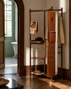 A tall wooden entryway organizer with integrated shoe storage and hanging rails, styled in a warm, sunlit room with wood floors and an arched doorway leading to a bathroom.