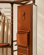 A close-up of a mid-century modern wardrobe organizer with a warm brown wood frame and cognac leather details, including a hanging pocket, a coat rack, and a mirrored drawer front.