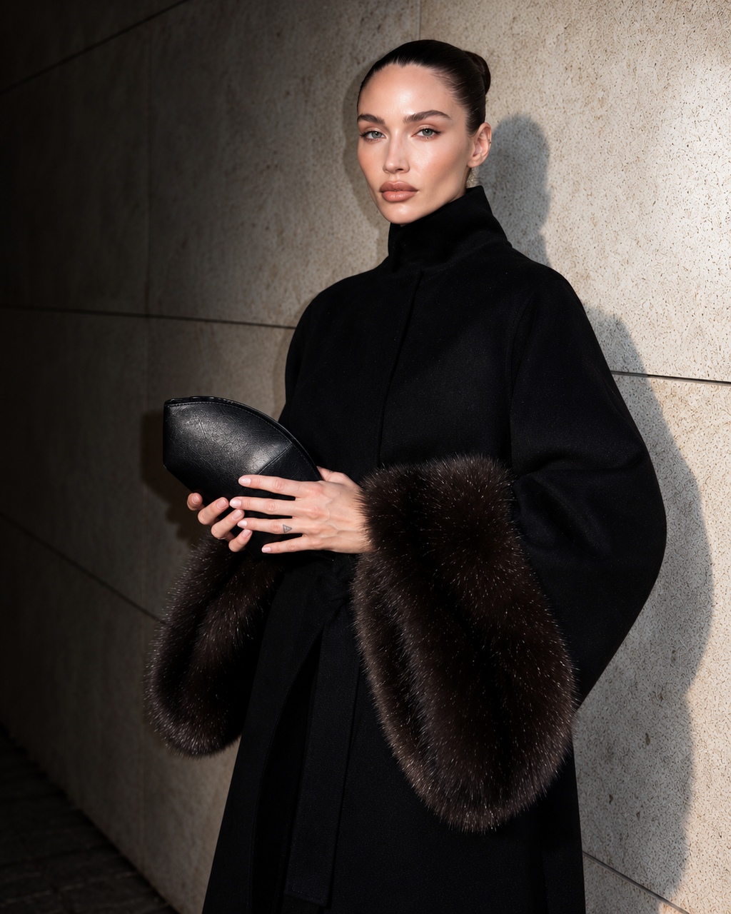 A model wearing a black high-neck coat with dark brown fur trim on the sleeves, holding a black leather clutch against a textured stone wall.