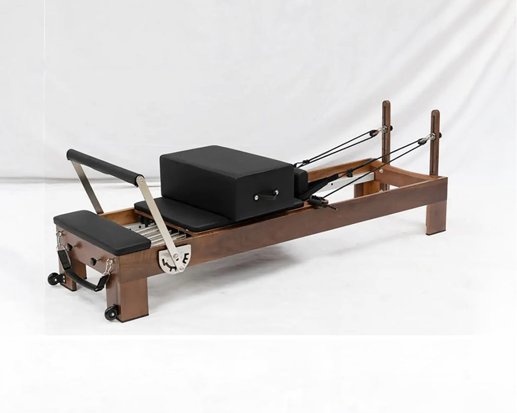 A wooden Pilates reformer machine with black foam padding, a black box, and metal springs, photographed on a white studio background.