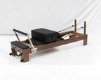 A wooden Pilates reformer machine with black foam padding, a black box, and metal springs, photographed on a white studio background.