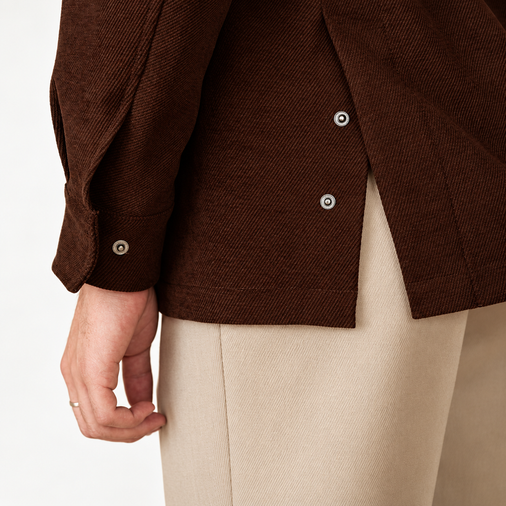 Close-up of a person wearing a dark brown textured jacket with silver buttons and light beige trousers, cropped to show the torso and hand against a plain light background.