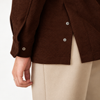 Close-up of a person wearing a dark brown textured jacket with silver buttons and light beige trousers, cropped to show the torso and hand against a plain light background.