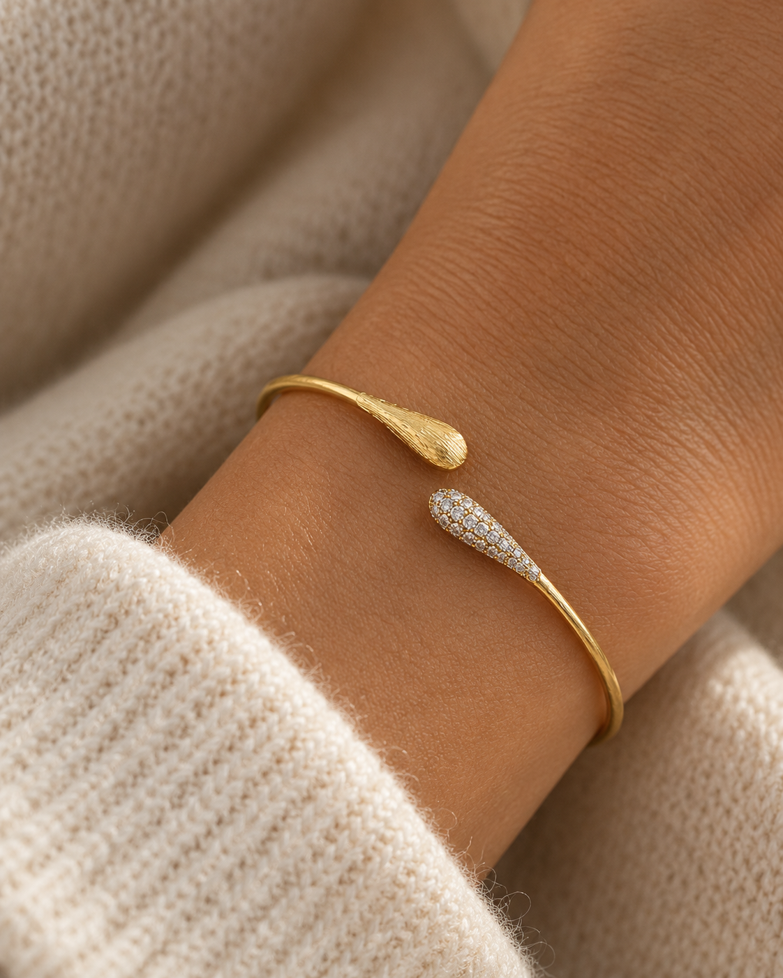 A close-up of a gold cuff bracelet with two teardrop-shaped ends, one textured gold and one pavé-set with small clear stones, worn on a wrist over a cream knit sweater.