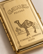 A close-up of a gold-toned Camel Filters cigarette case with an engraved camel and desert scene, plus the text 'Turkish & Domestic Blend'.