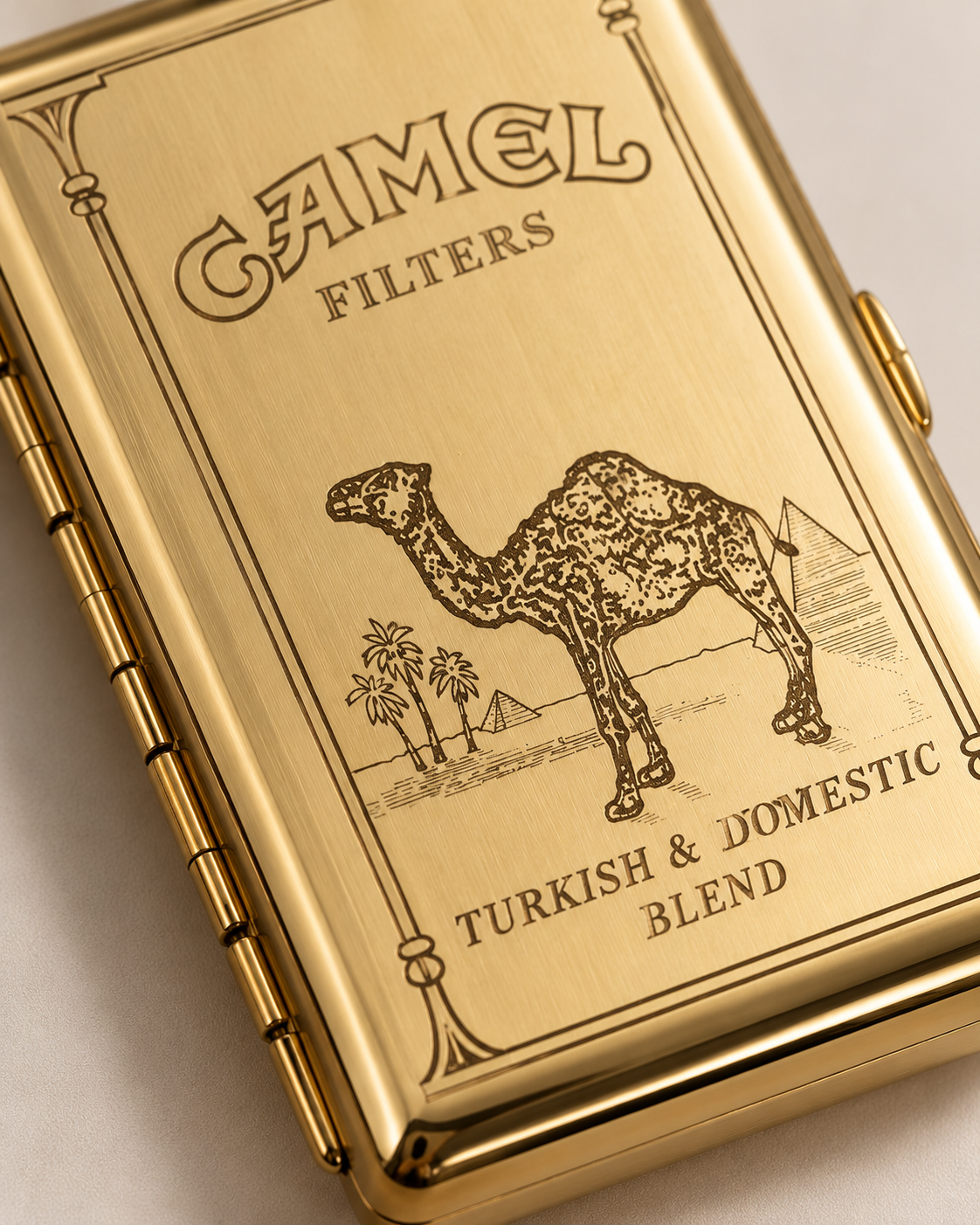 A close-up of a gold-toned Camel Filters cigarette case with an engraved camel and desert scene, plus the text 'Turkish & Domestic Blend'.