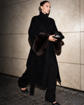 A model walks against a textured stone wall wearing a long black high-neck coat with dark brown fur trim on the collar and cuffs, paired with black trousers and holding a black clutch.