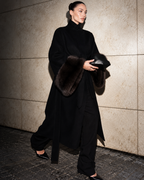 A model walks against a textured stone wall wearing a long black high-neck coat with dark brown fur trim on the collar and cuffs, paired with black trousers and holding a black clutch.