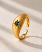 A gold crescent moon ring with a central green gemstone surrounded by a starburst pattern, set against a warm beige background with soft diagonal shadows.
