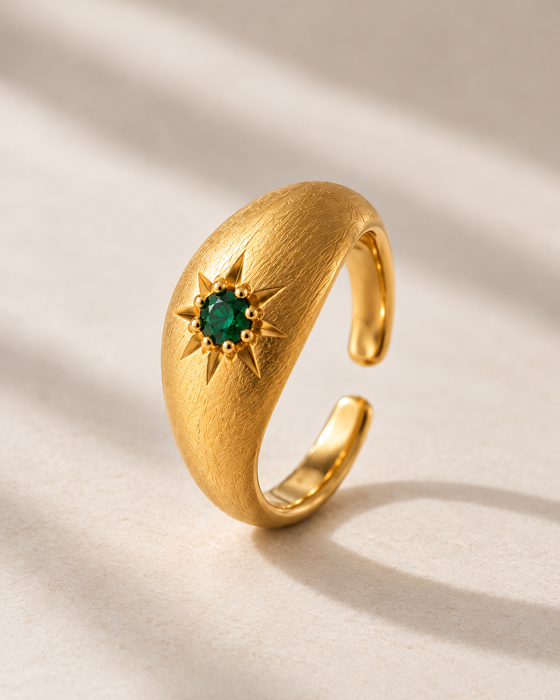 A gold crescent moon ring with a central green gemstone surrounded by a starburst pattern, set against a warm beige background with soft diagonal shadows.