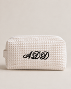 A cream-colored waffle-weave cosmetic pouch with a black embroidered monogram 'ADD' on the front, set against a solid light beige background.