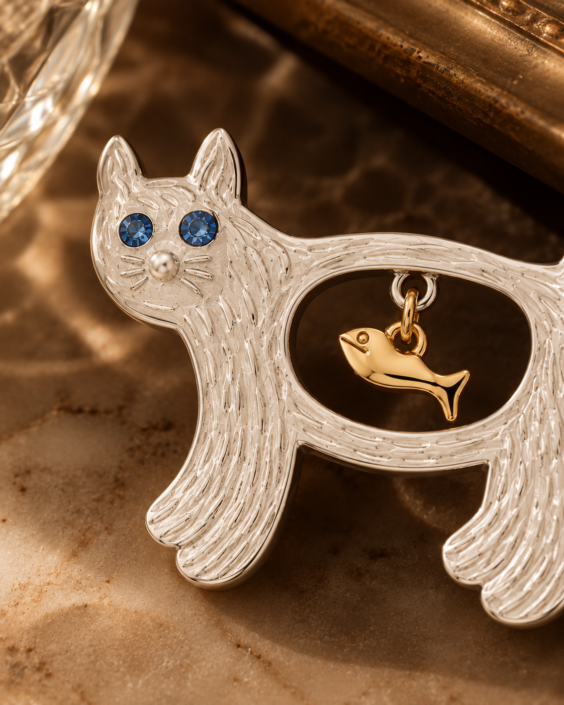 A silver cat-shaped pendant or brooch with textured detailing, featuring blue gemstone eyes, a small pearl-like nose, and a dangling gold fish charm inside the cat's body, shown on a warm brown surface.