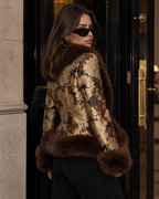 A woman wearing a metallic gold and brown patterned jacket with dark brown fur trim, black pants, and black sunglasses, standing in front of a black doorway.