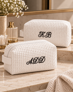 Two white waffle-weave cosmetic pouches with embroidered black monogram initials, displayed on a beige marble bathroom counter with a towel and toiletry items in the background.