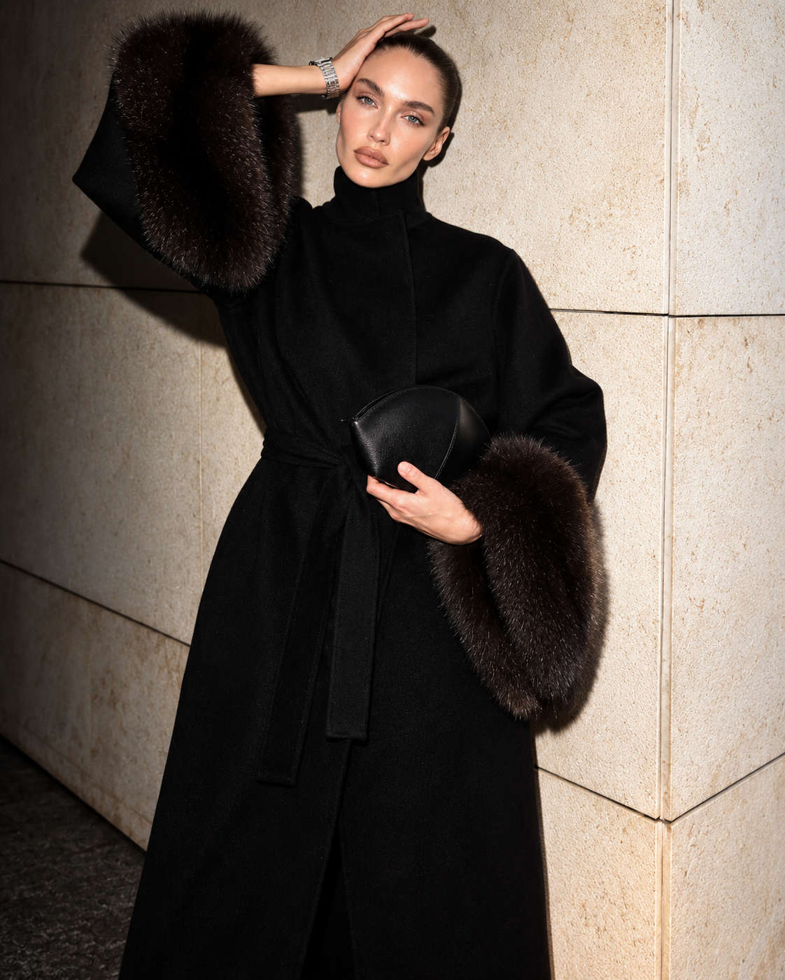 A model wearing a long black coat with luxurious dark brown fur trim on the sleeves and collar, holding a black leather clutch bag against a beige stone wall background.
