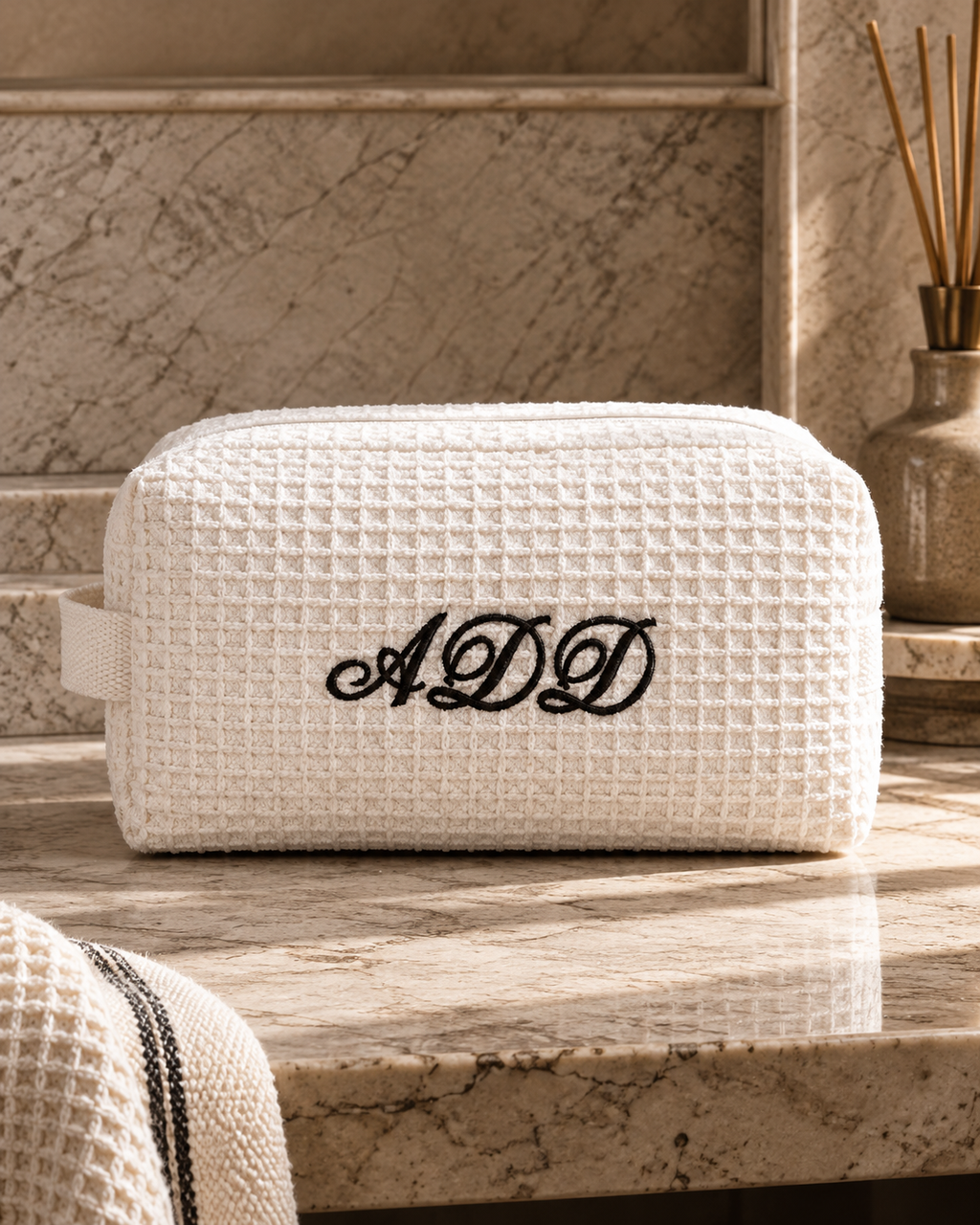 A white waffle-weave cosmetic pouch with black embroidered initials 'ADD' on the front, resting on a beige marble countertop in a bathroom setting.