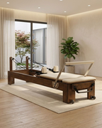 A wooden Pilates reformer machine with cream-colored padding and black springs, set on a light beige rug in a modern, sunlit room with a large window and indoor plant.