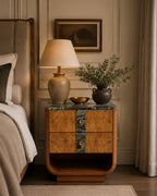 A styled bedroom scene featuring a vintage-style wooden nightstand with burl wood grain and a green marble top, styled with a table lamp, a small bowl, and a potted olive branch, next to a bed.