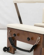 Close-up of a wooden exercise or therapy chair with cream upholstered seat and back, metal support frame, and black straps attached to the side.