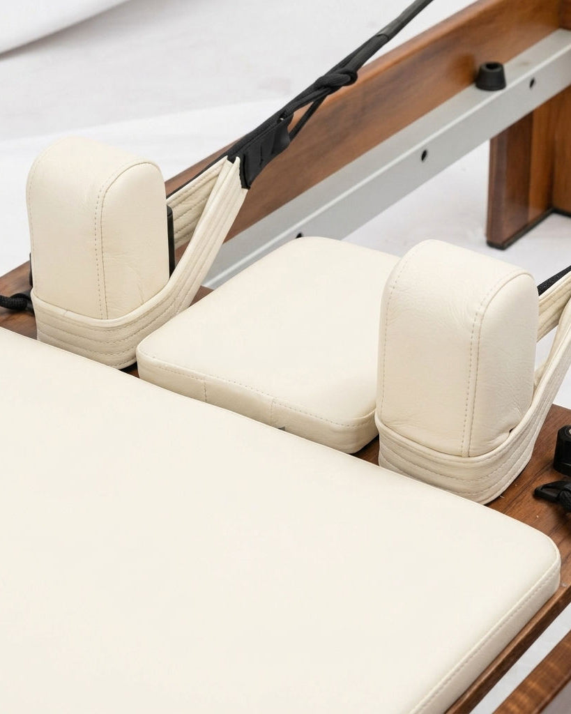 Close-up of a Pilates reformer machine showing cream-colored padded footboard, headrests, and straps over a wooden frame.
