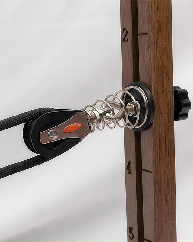 Close-up of a pulley system with a black rope, silver metal spring, and a wooden vertical rail marked with numbers, set against a light gray background.