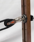 Close-up of a pulley system with a black rope, silver metal spring, and a wooden vertical rail marked with numbers, set against a light gray background.
