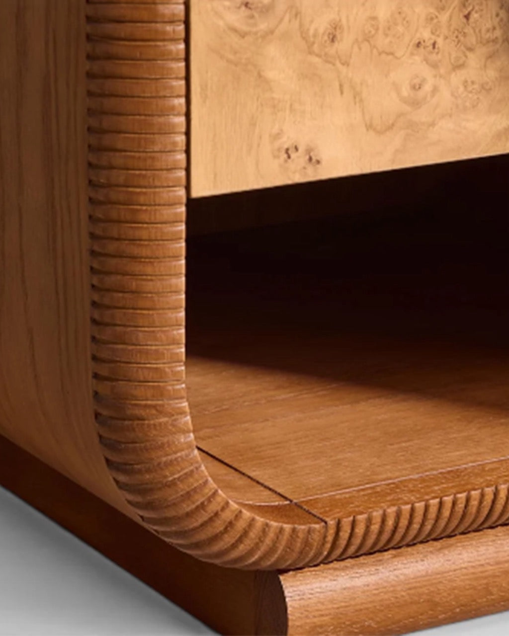 Close-up of a wooden cabinet corner showing a curved ribbed front panel, burl wood drawer front, and warm medium-brown wood shelf.