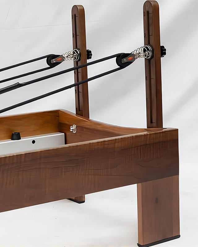 Close-up of a Pilates reformer machine showing the wooden frame, black resistance bands, pulleys with silver metal hardware, and part of the white weight stack on a white studio background.