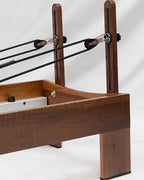 Close-up of a Pilates reformer machine showing the wooden frame, black resistance bands, pulleys with silver metal hardware, and part of the white weight stack on a white studio background.