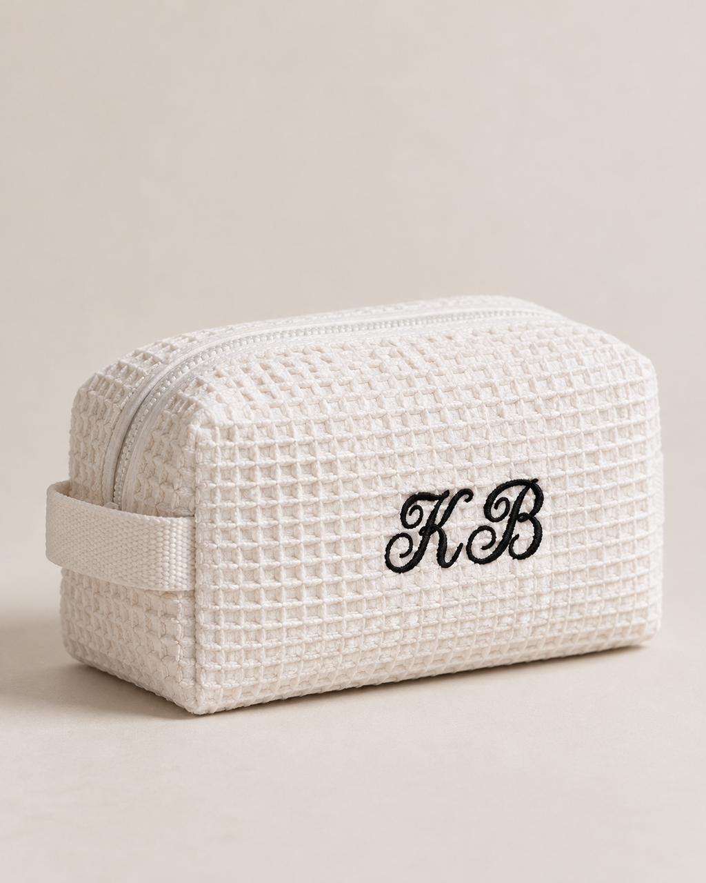 A cream-colored waffle-weave cosmetic pouch with a matching fabric loop handle and black embroidered monogram initials 'KB' on the front, set against a solid light beige background.