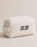 A cream-colored waffle-weave cosmetic pouch with a matching fabric loop handle and black embroidered monogram initials 'KB' on the front, set against a solid light beige background.