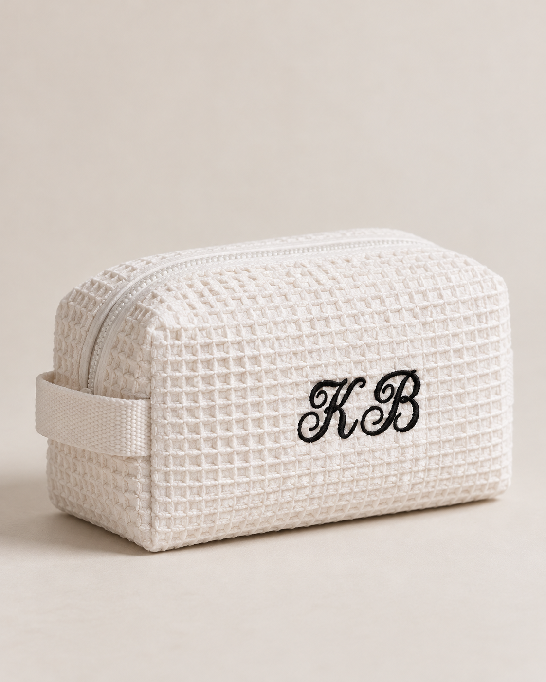 A cream-colored waffle-weave cosmetic pouch with a matching fabric loop handle and black embroidered monogram initials 'KB' on the front, set against a solid light beige background.