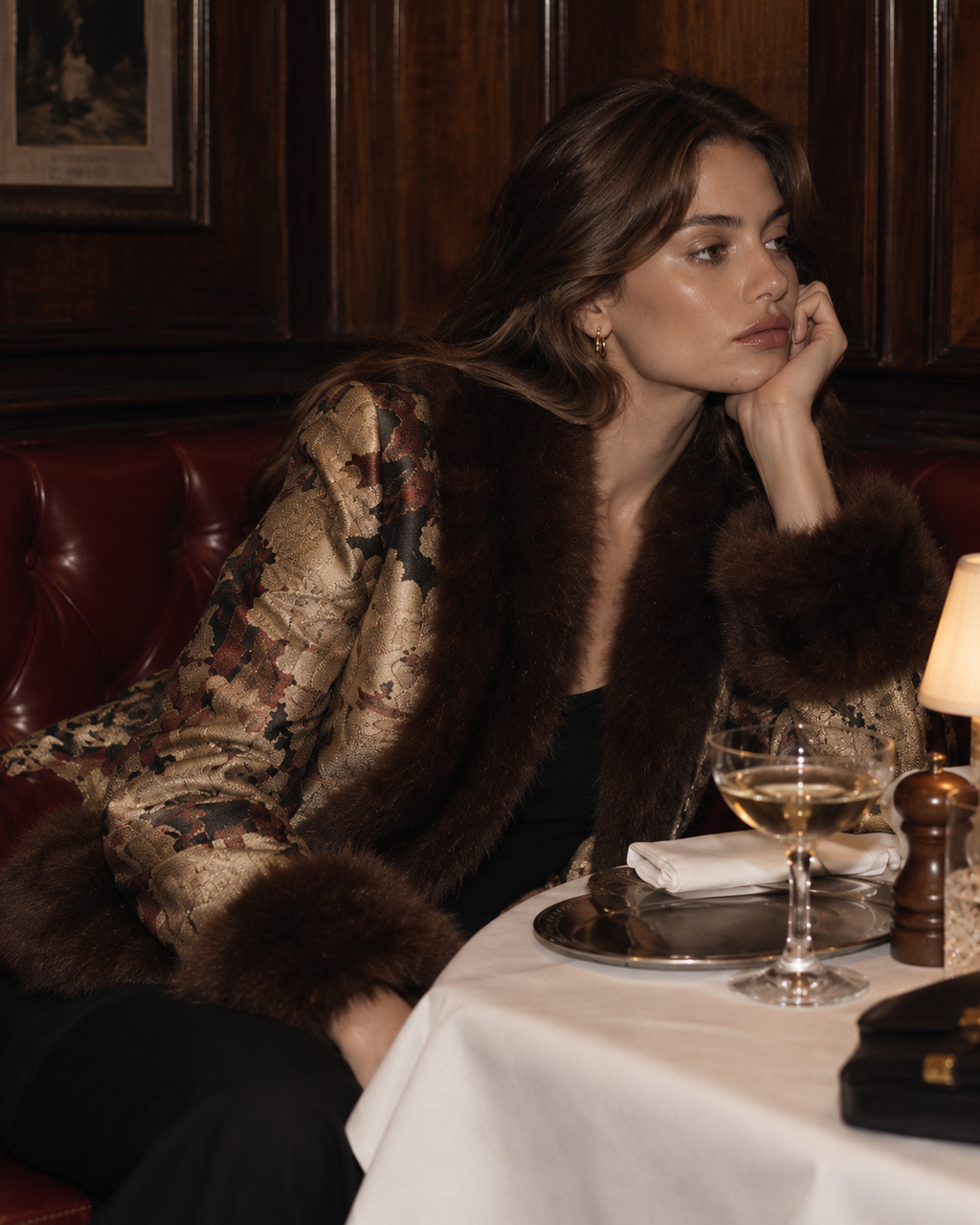 A woman in a luxurious dark brown fur-trimmed jacket with a gold-toned patterned body sits in a dimly lit restaurant booth next to a table with a glass of white wine.