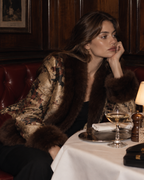 A woman in a luxurious dark brown fur-trimmed jacket with a gold-toned patterned body sits in a dimly lit restaurant booth next to a table with a glass of white wine.