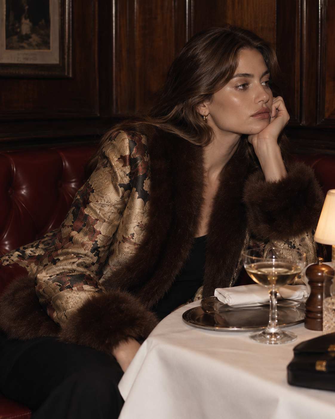 A woman in a luxurious dark brown fur-trimmed jacket with a gold-toned patterned body sits in a dimly lit restaurant booth next to a table with a glass of white wine.