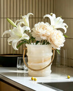 White lilies and cream peonies arranged in a ribbed translucent cream vase with a gold metal handle, set on a kitchen countertop against a beige tiled wall.