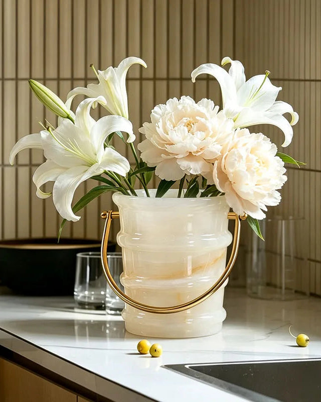 White lilies and cream peonies arranged in a ribbed translucent cream vase with a gold metal handle, set on a kitchen countertop against a beige tiled wall.