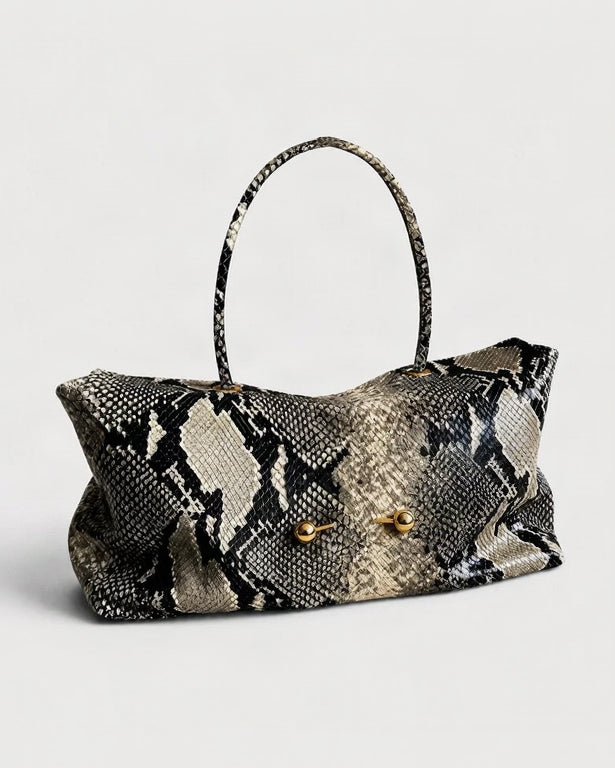A snakeskin-patterned handbag with a black, beige, and gray scale design, featuring a single top handle and two gold-tone metal accents on the front.