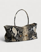 A snakeskin-patterned handbag with a black, beige, and gray scale design, featuring a single top handle and two gold-tone metal accents on the front.