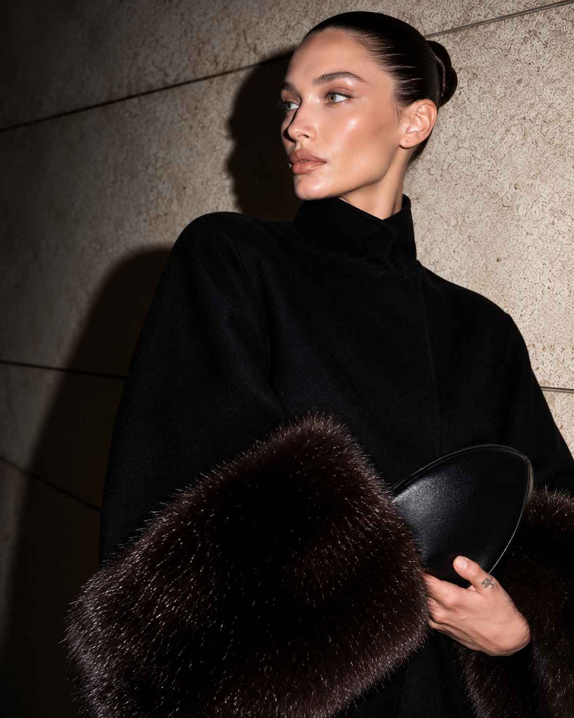 A model in a black high-neck coat holds a black hat with a dark brown fur trim against a textured stone wall background.