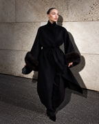 A model walks against a textured stone wall wearing a long black coat with dark fur trim on the sleeves and a matching waist tie.