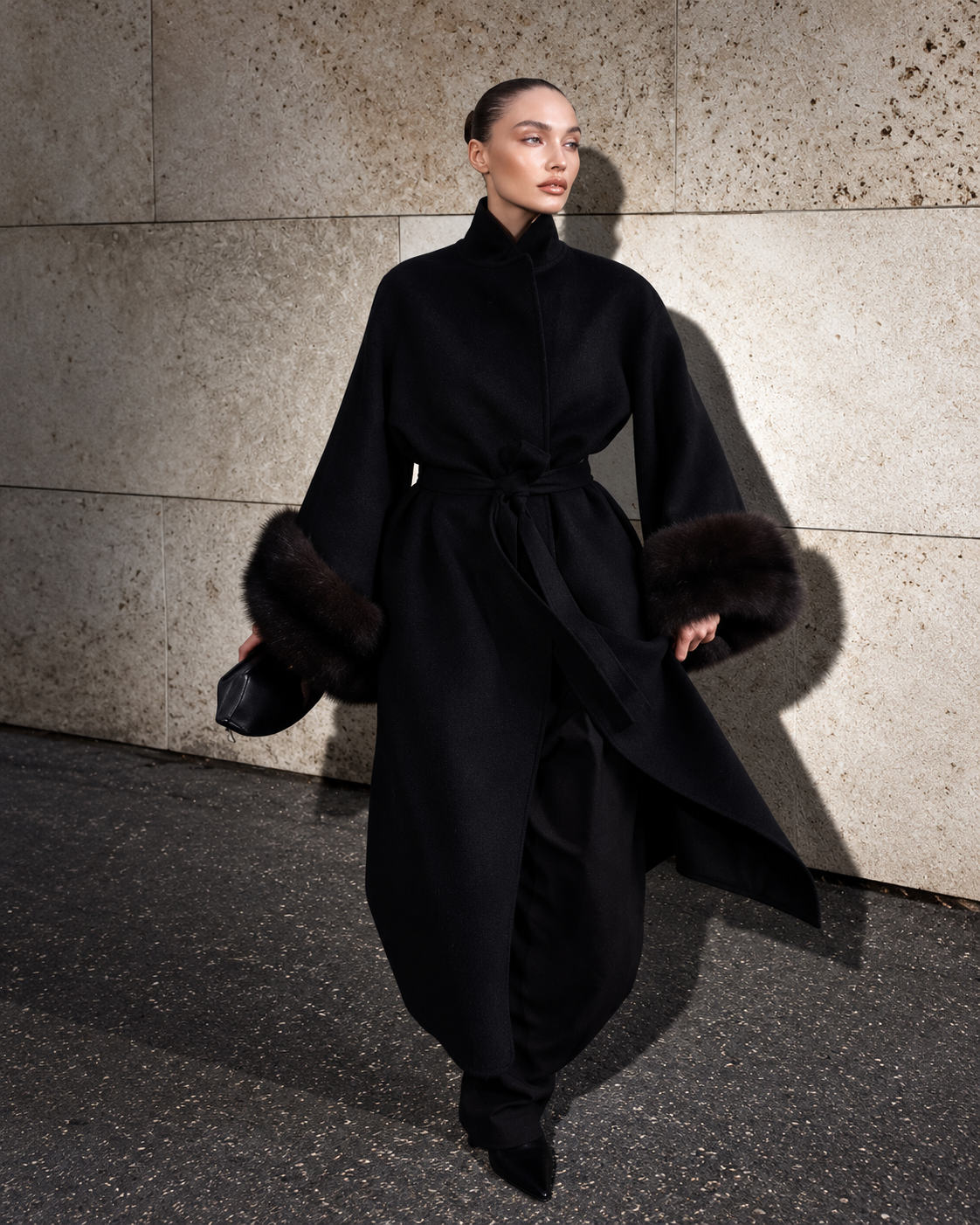 A model walks against a textured stone wall wearing a long black coat with dark fur trim on the sleeves and a matching waist tie.