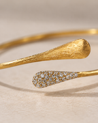Gold open cuff bangle bracelet with a textured brushed gold end and a pavé-set diamond accent end, photographed on a neutral beige background.
