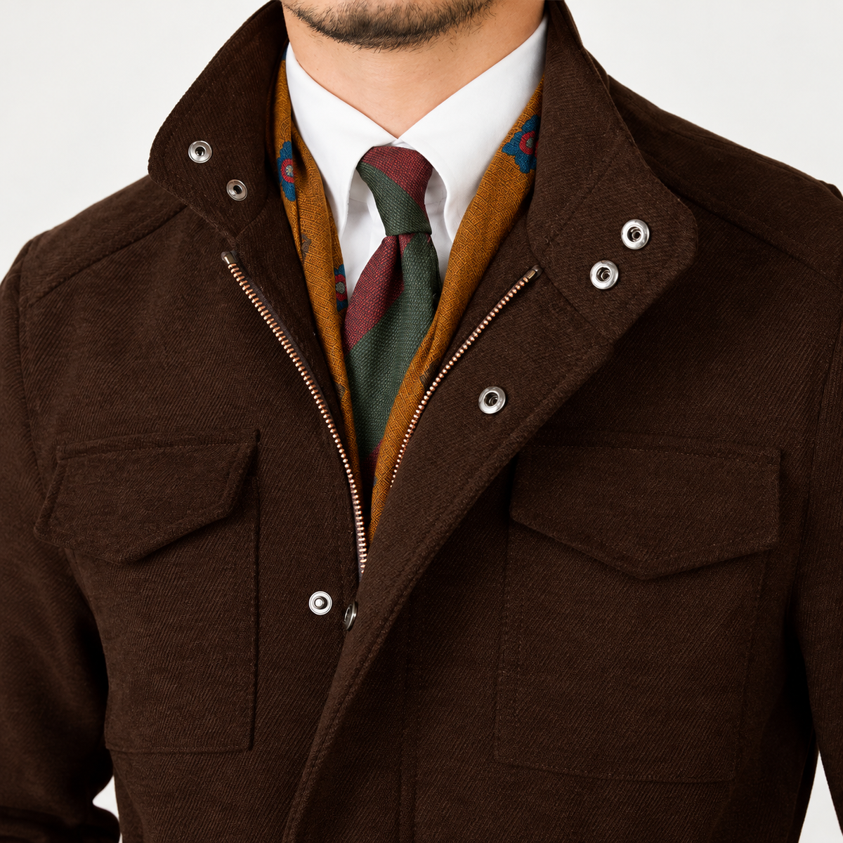 A close-up of a man wearing a dark brown textured jacket with silver snap buttons and a zipper, layered over a white dress shirt, a rust-orange patterned scarf, and a green and burgundy tie.