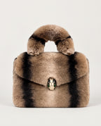 A structured faux fur handbag with a rounded top handle, featuring alternating vertical stripes of light brown, black, and beige, and a snake-head clasp at the center.